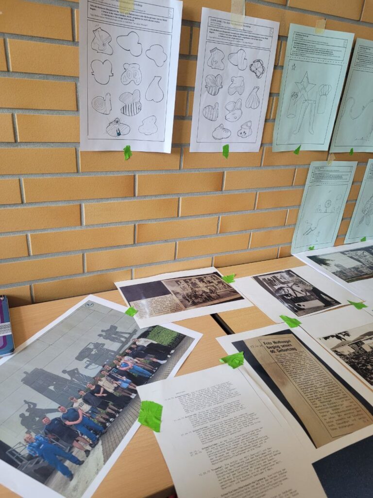 Das Foto zeigt einen Tisch in einer Schule, der vor einer Wand steht. An dieser Wand sind Arbeitsblätter befestigt, auf dem Tisch liegen verschiedene Fotos und historische Aufnahmen.