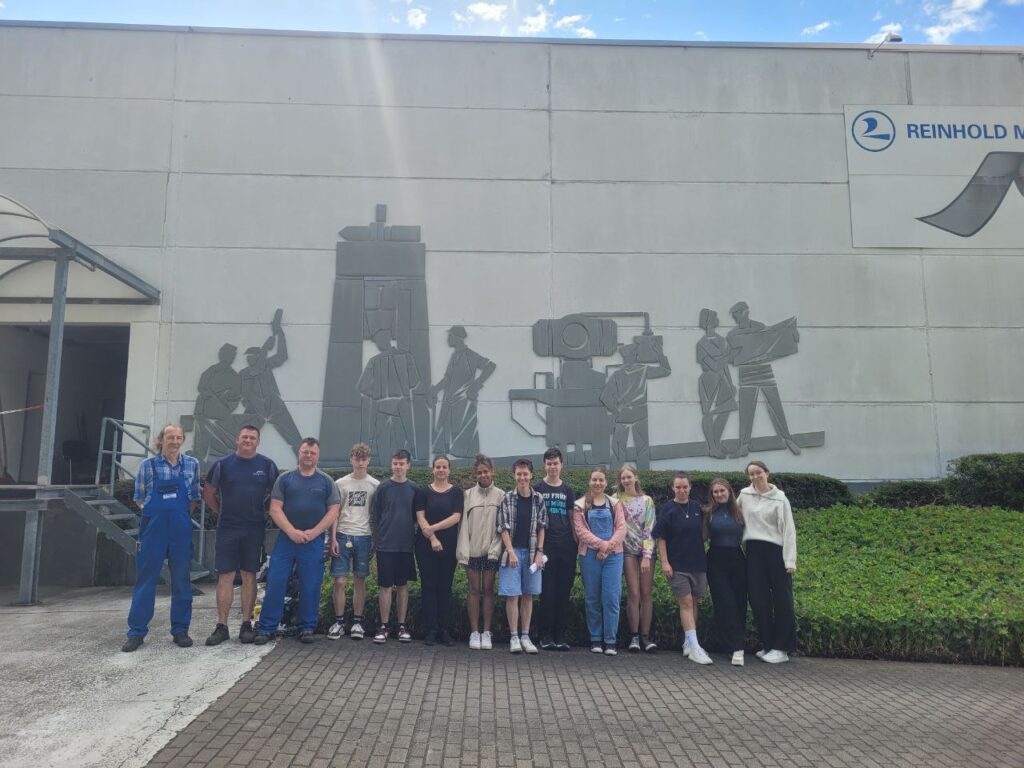 Das Foto zeigt eine Gruppe Schülerinnen und Schüler mit mehreren ännern in Arbeitskleidung. Die Gruppe steht vor einem Industriegebäude, an dessen Fassade sich ein Wandkunstwerk aus Aluminium befindet.