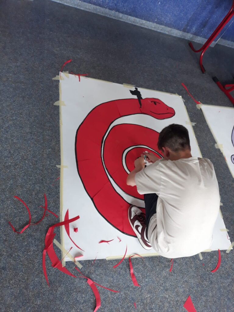 Das Foto zeigt einen Jungen von hinten. Dieser hockt über einem gro0en Plakat und schneidet etwas aus buntem Papier aus.