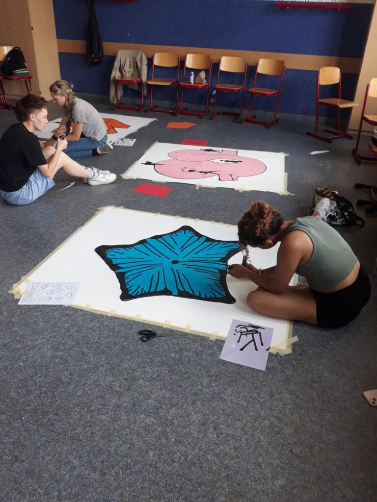 Das Foto zeigt einen Klassenraum. Auf dem Boden sitzen drei junge Menschen, die an einem vor sich liegenden Kunstwerk auf einem großen Plakat arbeiten.